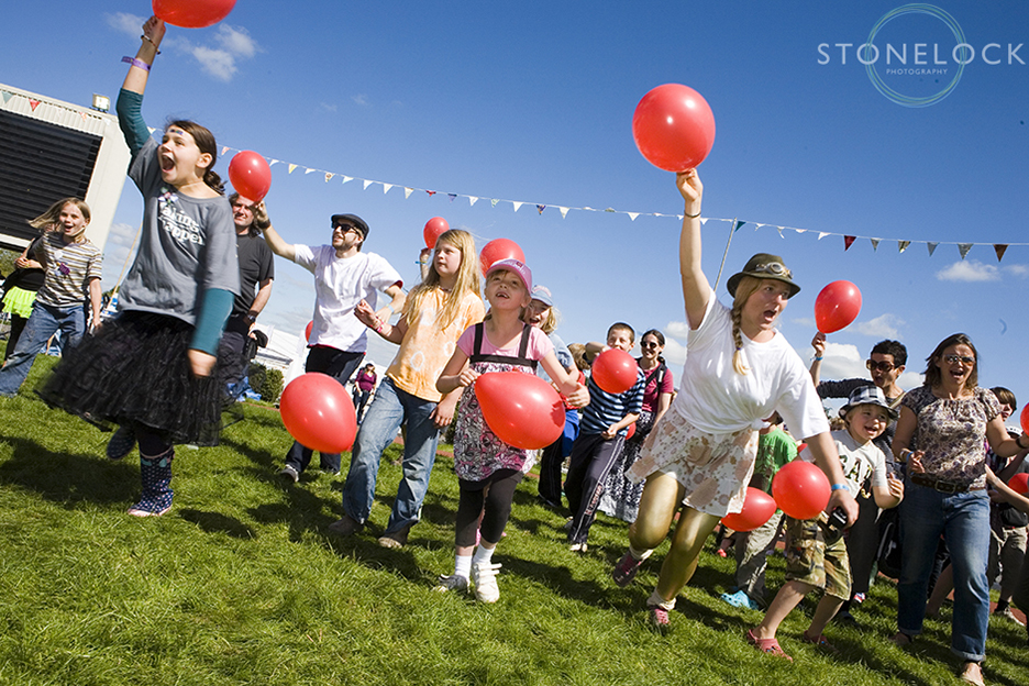 Balloon games in the Arena at Cheltenham Racecourse during Greenbelt Arts Festival, a group of participants run towards the camera holding bright red gallons that stand out well against the blue sky