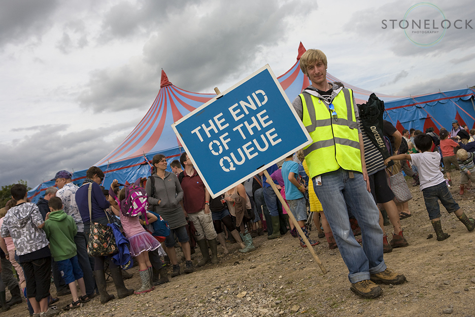 A steward stands outside a big top tent at Greenbelt Arts Festival holding a sign that says the end of the queue