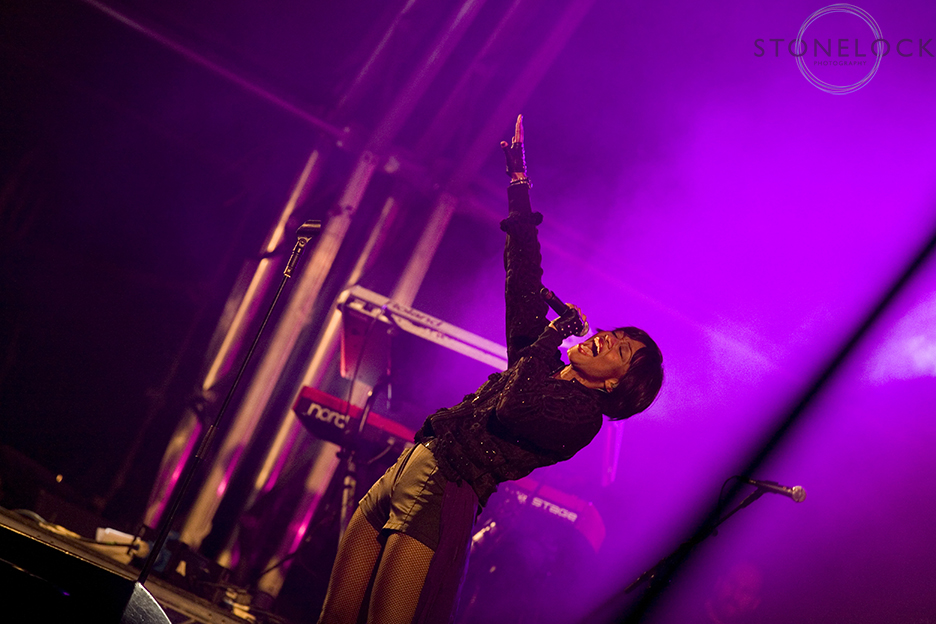Beverley Knight performs on MainStage at Greenbelt Arts Festival. Her arm is up in the air as she stands in front of a bright pink light
