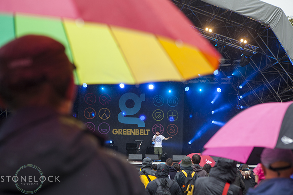 A man stands in the rain with a large rainbow umbrella watching Hobit perform on MainStage at Greenbelt Arts Festival. Shot from behind the man and his umbrella frame the artiste.