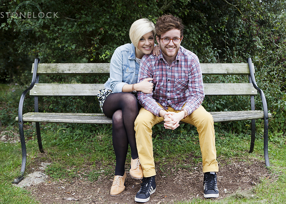 The couple sit on a bench on the Downs in Bristol during their engagement shoot, the girl has her arms interlinking with the guy