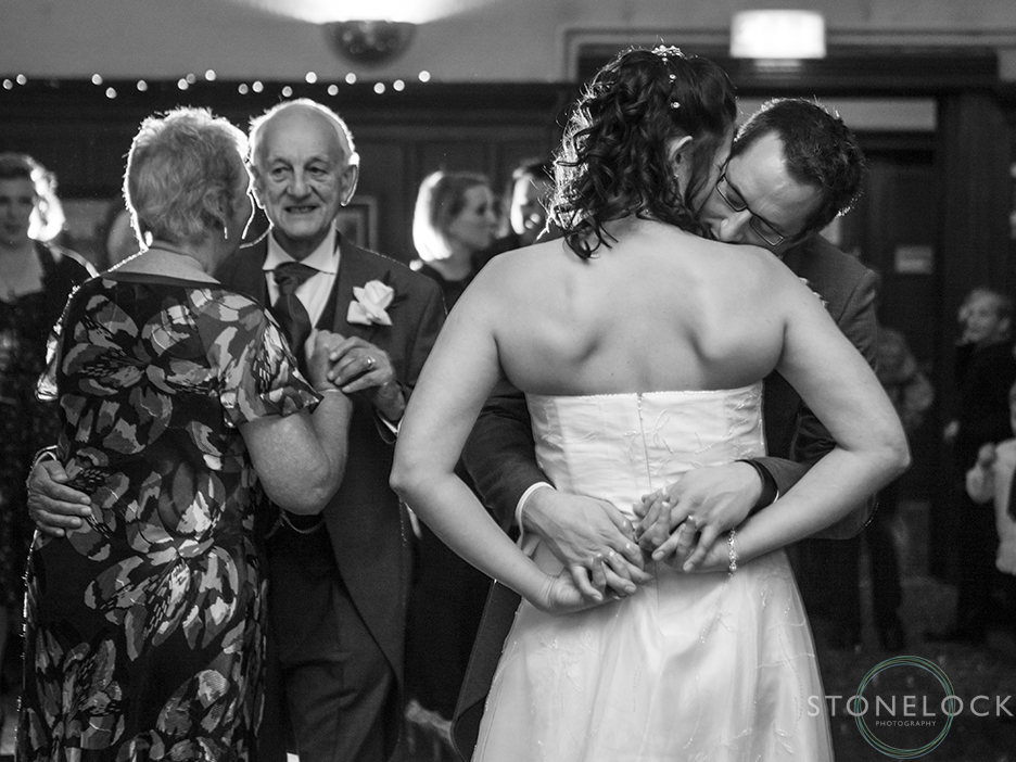 A bride and groom during their first dance, he leans over her shoulder and rests his head, in the background the mother of the bride and father of the bride are dancing, the photo is in black and white