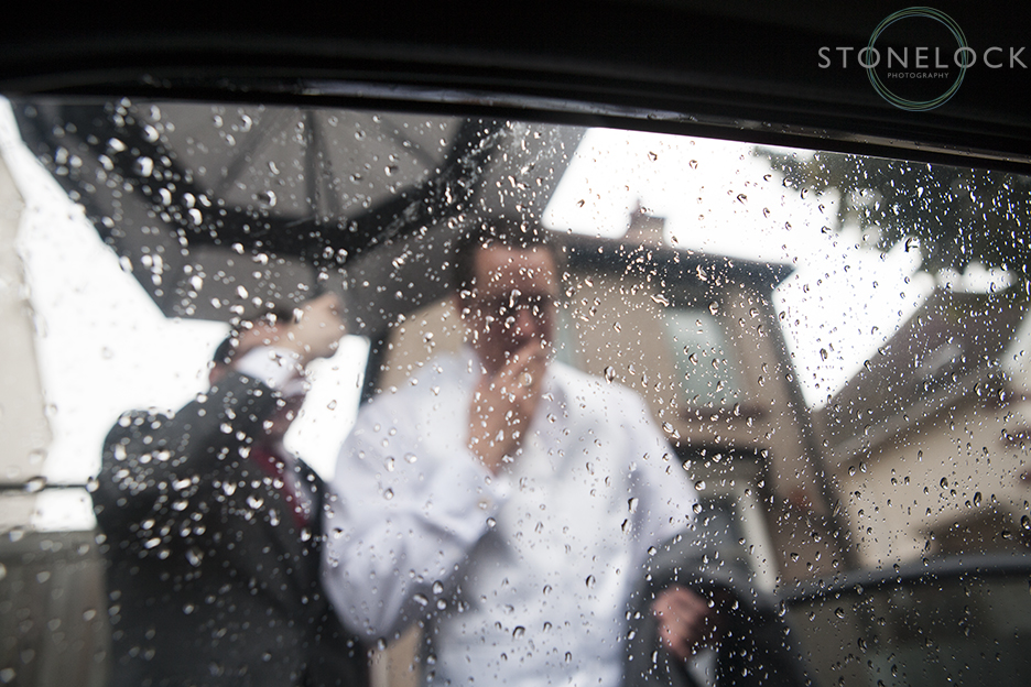 The groom smokes a last cigarette before the wedding, it is raining and he is photographed from behind a window covered in raindrops giving an abstract and distorted feel to the photo