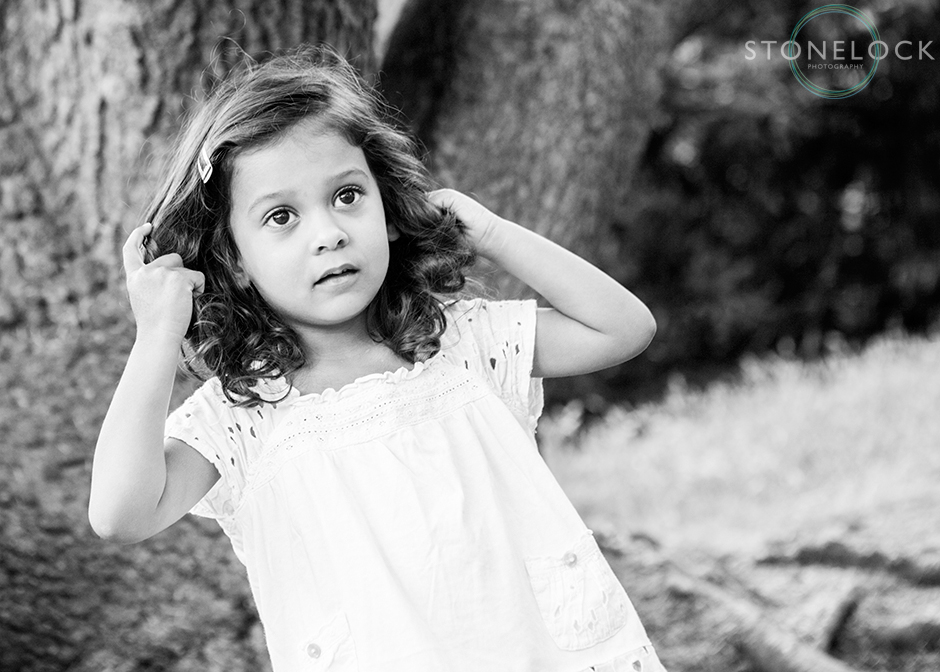 A young girl stands by a tree and pulls her hair off of her face, a very natural and informal portrait