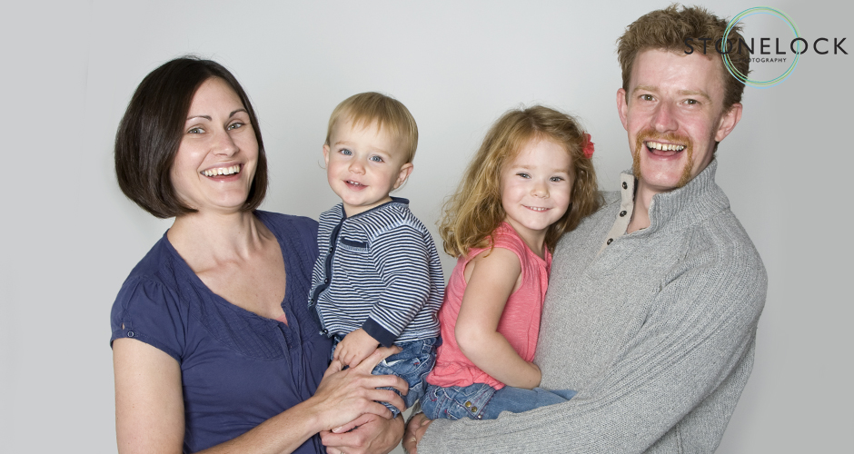 A family of four pose in the studio, Mum is holding the one year old boy and Dad is holding the four year old girl