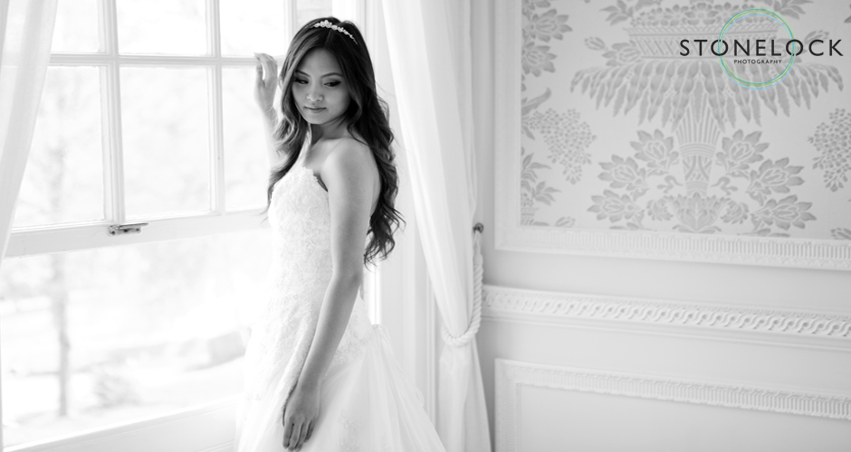 A bride, standing by a window peers over her should and back onto the room, showing off the full effect of her dress