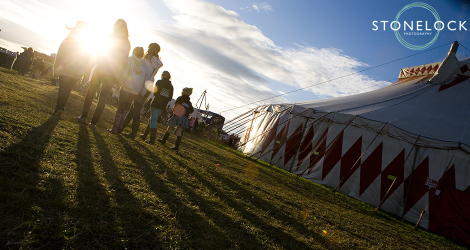 A group of people walk away from the camera into the sunsetting at Greenbelt Festival. A big top tent sits to the right hand side.