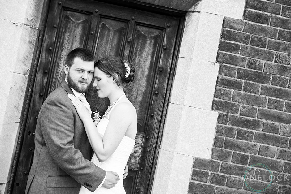 The bride and groom pose outside a wooden door, the photos is cropped at an angle and the groom rests his head on the brides head